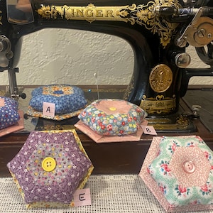May include: An antique Singer sewing machine with gold accents and a collection of handmade pin cushions. The pin cushions are in various shapes and patterns, including floral and geometric designs, with a button in the center of each.