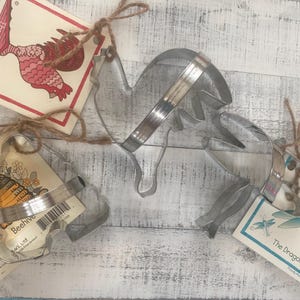 May include: Three silver metal cookie cutters in the shapes of a rooster, a beehive, and a dragonfly. Each cutter is attached to a square card with a corresponding illustration and the text The Rooster, Beehive, or The Dragonfly.