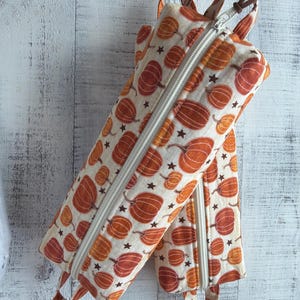 May include: Two rectangular fabric pencil cases featuring a repeating pattern of orange pumpkins and small dark brown stars on a cream background. Each case has a white zipper closure and a brown leather cord loop attached to the top end.