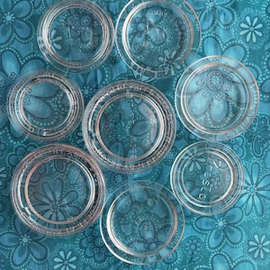 May include: A collection of seven clear glass circular lids of varying sizes, each featuring a raised rim and embossed text. The lids are arranged in a cluster, showcasing their transparent, smooth surfaces and distinct circular designs.