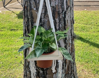 May include: A white macrame plant hanger with a wooden shelf and a potted plant hanging from a tree.