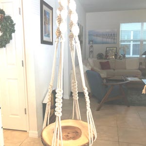 May include: A macrame plant hanger with a natural wood slice base. The off-white rope is knotted and woven, creating a decorative design. The wood slice has a natural bark edge and a warm, brown color.