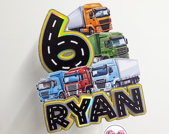 Może przedstawiać: Topper na tort z numerem 6 z wzorem drogi, imieniem "RYAN" w tym samym wzorze drogi i czterema zabawkowymi ciężarówkami. Topper jest na przezroczystym plastikowym patyku z różowo-białym okrągłym naklejką z napisem "Frankie & Me".