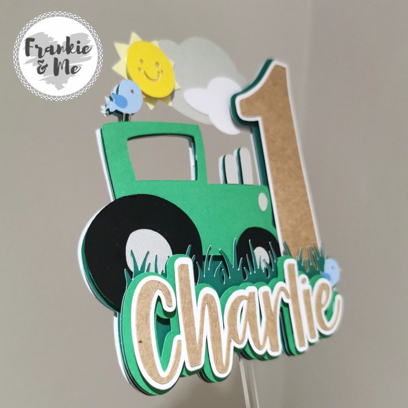 K&ouml;nnte beinhalten: Gr&uuml;ner Traktor-Kuchentopper mit einer braunen Nummer eins, einer gelben Sonne, blauen V&ouml;geln und dem Namen Charlie in braunen Buchstaben.