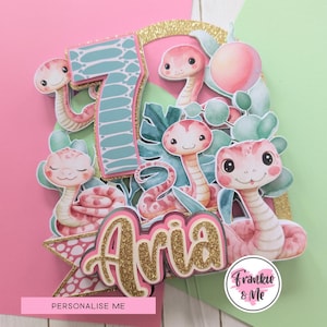 May include: A personalised cake topper for a seventh birthday, featuring cartoon snakes, the name 'Aria', and tropical foliage. The design uses pink, gold, and teal colours. The text 'Personalise Me' is included, along with the Frankie & Me logo.