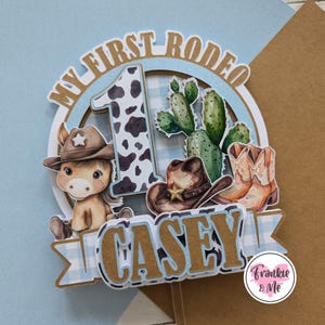 Pu&ograve; includere: Un topper per torta blu e marrone con il testo "My First Rodeo" e un cappello da cowboy, un cactus e un numero uno con una stampa di mucca. Il nome "Casey" &egrave; scritto in lettere marroni sul topper per torta.