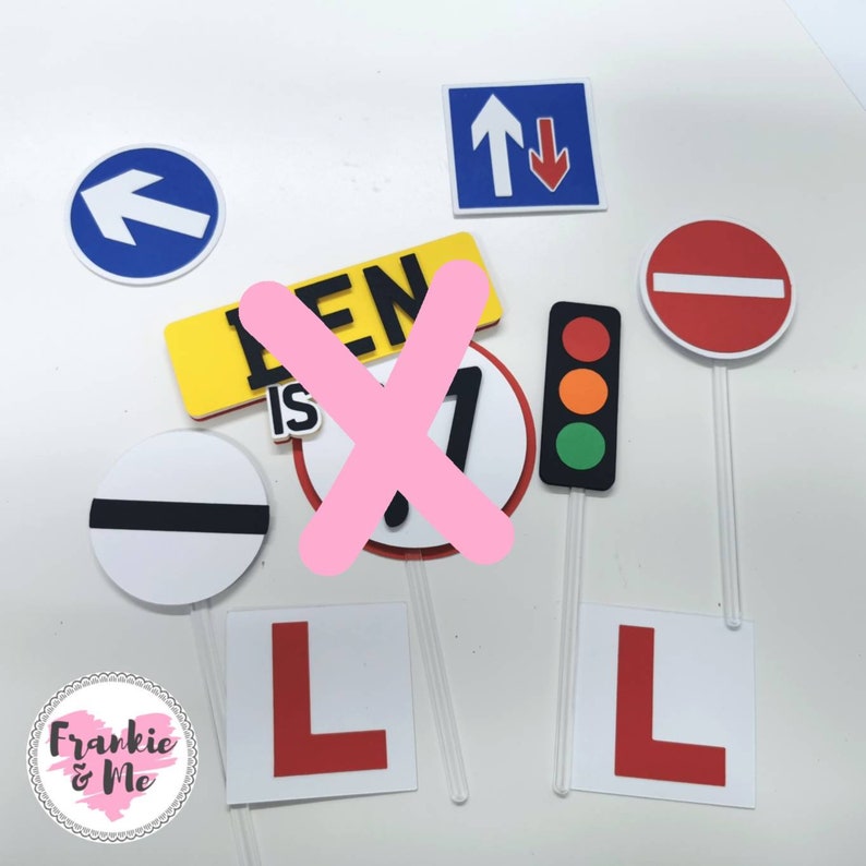 Puede incluir: Un conjunto de ocho adornos para pasteles con diferentes se&ntilde;ales de tr&aacute;fico, incluyendo una se&ntilde;al de stop, una se&ntilde;al de ceder el paso, una flecha de giro a la izquierda, un sem&aacute;foro y una se&ntilde;al de conductor novel. Los adornos est&aacute;n hechos de espuma y tienen un fondo blanco con detalles rojos, negros y amarillos. El texto "LEN IS 1" es visible en uno de los adornos.