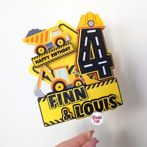 May include: Yellow and black construction themed cake topper with the number 4, a dumper truck, an excavator, and the names Finn & Louis. The topper says "Happy Birthday".