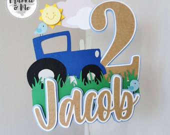 May include: A blue tractor cake topper with a smiling sun, clouds, and two blue birds. The topper also includes the number 2 and the name Jacob in brown letters.