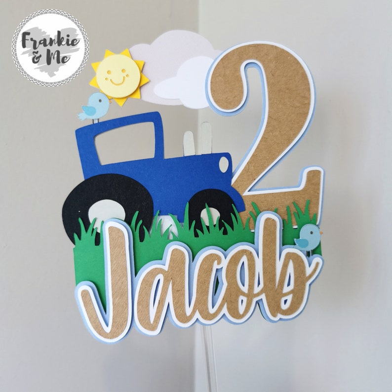 K&ouml;nnte beinhalten: Ein blauer Traktor-Kuchentopper mit einer l&auml;chelnden Sonne, Wolken und zwei blauen V&ouml;geln. Der Topper enth&auml;lt auch die Zahl 2 und den Namen Jacob in braunen Buchstaben.