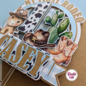 Pu&ograve; includere: Un topper per torta blu e marrone con il nome "Casey" e le parole "My First Rodeo". Il topper presenta un cappello da cowboy, stivali da cowboy, un cactus e una stampa di mucca.