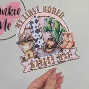 Pu&ograve; includere: Un topper per torta rosa e marrone con un numero uno con stampa di mucca, un cavallo di cartone animato con un cappello da cowboy, un cactus e il testo "My First Rodeo" e "Kayley Rae".