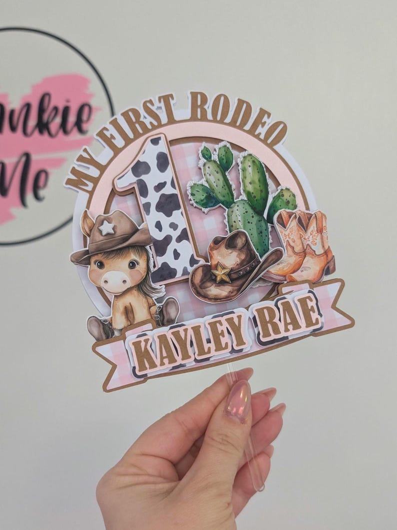 Pu&ograve; includere: Un topper per torta rosa e marrone con un numero uno con stampa di mucca, un cavallo di cartone animato con un cappello da cowboy, un cactus e il testo "My First Rodeo" e "Kayley Rae".