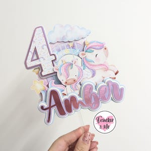 May include: A pink and purple unicorn cake topper with a sparkly number 4, a cloud, and the name "Amber" in pink and purple glitter. The topper is on a clear plastic stick and has a small pink circle with the text "Frankie & Me" at the bottom.