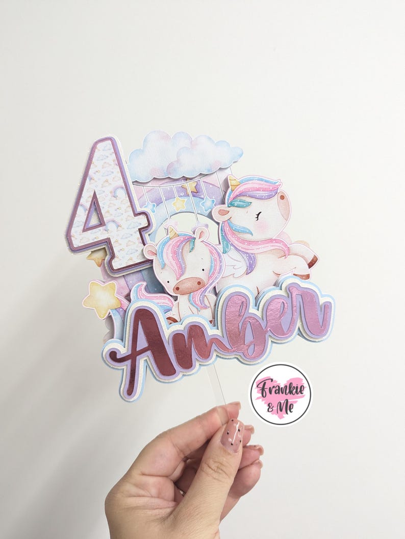 May include: A pink and purple unicorn cake topper with a sparkly number 4, a cloud, and the name "Amber" in pink and purple glitter. The topper is on a clear plastic stick and has a small pink circle with the text "Frankie & Me" at the bottom.
