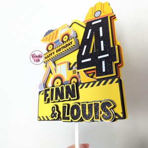 May include: Yellow and black construction themed cake topper with the number 4, two dumper trucks, and the names Finn & Louis. The topper is on a white stick.