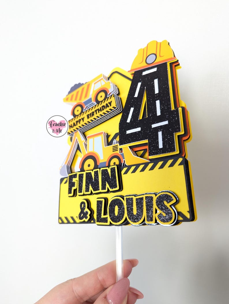 May include: Yellow and black construction themed cake topper with the number 4, two dumper trucks, and the names Finn & Louis. The topper is on a white stick.