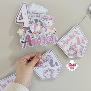 May include: A purple and pink unicorn themed birthday banner with the text "Amber is 4". The banner is made of cardstock and features a unicorn with a rainbow mane and tail. The banner is strung together with twine.