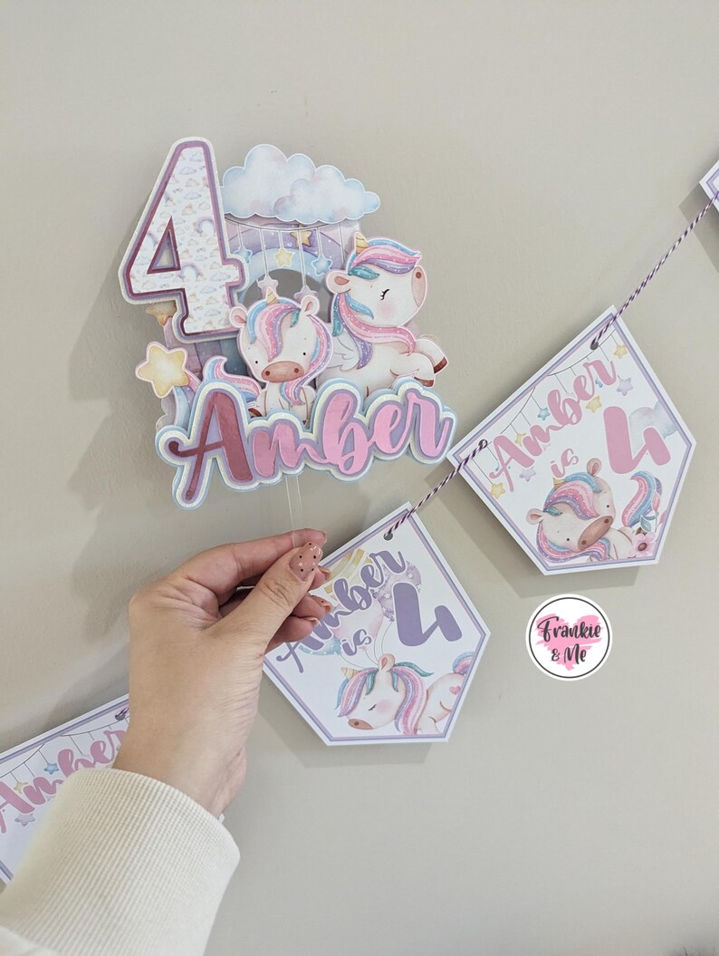 May include: A purple and pink unicorn themed birthday banner with the text "Amber is 4". The banner is made of cardstock and features a unicorn with a rainbow mane and tail. The banner is strung together with twine.