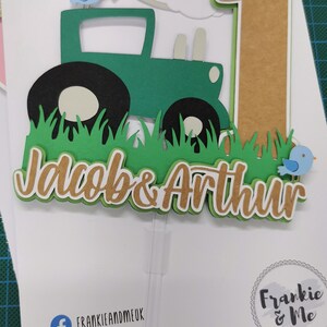 K&ouml;nnte beinhalten: Gr&uuml;ner Traktor-Kuchentopper mit einer l&auml;chelnden Sonne, zwei blauen V&ouml;geln und dem Text "Jacob & Arthur" in braunen Buchstaben. Die Nummer eins ist aus braunem Papier mit gr&uuml;nem Umriss.