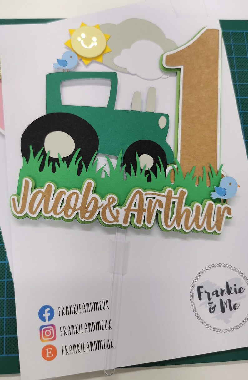 K&ouml;nnte beinhalten: Gr&uuml;ner Traktor-Kuchentopper mit einer l&auml;chelnden Sonne, zwei blauen V&ouml;geln und dem Text "Jacob & Arthur" in braunen Buchstaben. Die Nummer eins ist aus braunem Papier mit gr&uuml;nem Umriss.