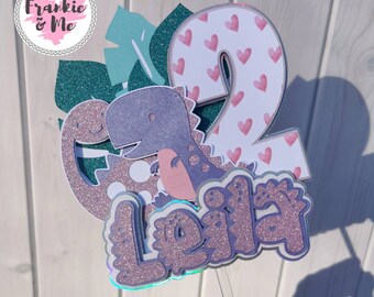 May include: A glittery cake topper featuring a purple dinosaur, the name "Leila", and the number "2" decorated with pink hearts. The topper includes green leaves and the text "Frankie & Me".