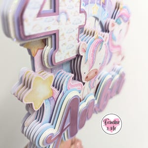 May include: A multi-layered cake topper featuring a unicorn, a star, and the number 4. The topper is decorated with pastel colors and glitter. The name "Ada" is written in pink glitter on the topper. The topper is made by Frankie & Me.