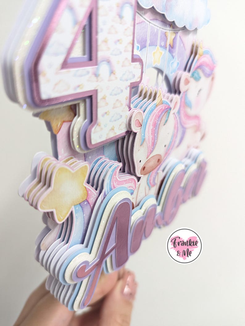 May include: A multi-layered cake topper featuring a unicorn, a star, and the number 4. The topper is decorated with pastel colors and glitter. The name "Ada" is written in pink glitter on the topper. The topper is made by Frankie & Me.