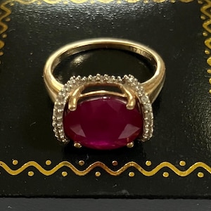 May include: A gold ring featuring a large, oval-cut, deep red gemstone set in the center. The setting is surrounded by a halo of small, clear, sparkling stones. The band is smooth and polished gold, with a visible hallmark stamped on the inner side.