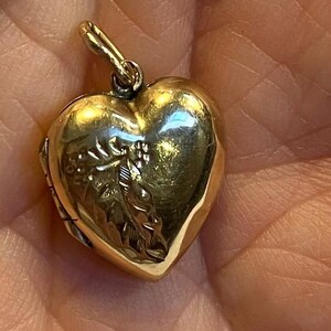 May include: A gold-toned heart-shaped locket pendant featuring an engraved leaf pattern on the front surface. The locket has a small metal loop at the top for attachment to a chain or necklace, with a polished finish that reflects light.