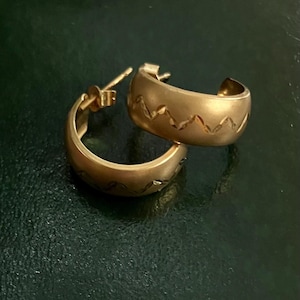 Peut inclure: Une paire de créoles en or avec une finition mate et un motif en zigzag répété gravé au centre de l'anneau. Chaque boucle d'oreille comprend un mécanisme de fermeture à tige, présentant un design incurvé en demi-cercle.