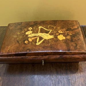 May include: A rectangular wooden music box with a curved lid. The box is a dark brown with a wood grain pattern. The lid features an inlaid design of a harp, musical notes, and flowers in gold and cream colors. A small keyhole is visible.