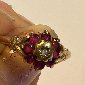 Peut inclure: Une bague en or avec une pierre centrale claire entourée d'un groupe de pierres circulaires rose foncé disposées en motif floral. L'anneau en métal présente des gravures décoratives en forme de feuilles sur les côtés, maintenant le sertissage.