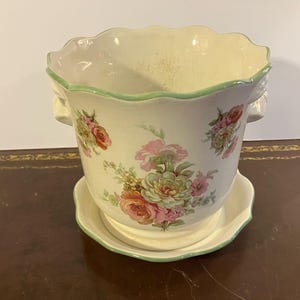 Könnte beinhalten: Ein weißer Keramiktopf mit grünem Rand und Blumenmuster. Der Topf hat zwei Griffe an den Seiten und wird mit einer passenden Untertasse geliefert.