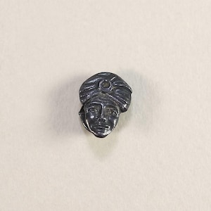 May include: A small, silver-coloured metal sculpture of a human face wearing a turban. The face has detailed features, including eyes, a nose, and a mouth. The turban has textured folds and a circular detail at the centre. The sculpture is likely a decorative item or jewellery component.