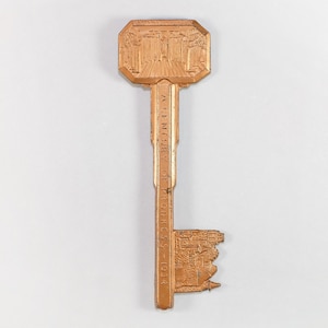 May include: A copper-colored key with an ornate design. The key's head features a building facade, and the shaft reads "A Century of Progress - 1934." The key's bit also has a building design. The key is likely a souvenir or commemorative item.