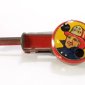 May include: A vintage red metal toy siren featuring a circular face with a yellow background and a graphic illustration of a firefighter wearing a helmet labeled F.D. CHIEF. The device includes a metal handle and a spring mechanism for operation.