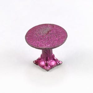 Può includere: Un piccolo tavolo rosa metallizzato con piano rotondo e base ornata. Il tavolo ha una superficie strutturata ed è probabilmente un mobile in miniatura o per casa delle bambole. La base ha una forma quadrata con piedini decorativi.
