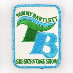 May include: Embroidered patch with a white background, blue border, and text. The patch features the words "TOMMY BARTLETT" in green and blue, along with a stylized "TB" in green and blue. The text "SKI-SKY-STAGE SHOW" is at the bottom.