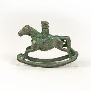 May include: A small, antique-style metal rocking horse figurine. The horse is ridden by a figure and has a weathered, green-toned patina. The base is curved, allowing the horse to rock. The figurine is a decorative item.