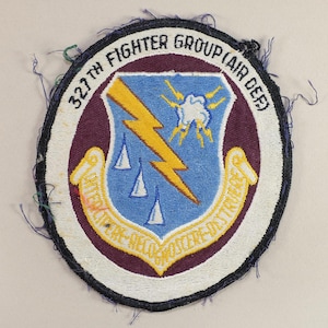 May include: A vintage embroidered patch with a maroon background and white border. The patch features a blue shield with a yellow lightning bolt and three white triangles. The text "327th Fighter Group Air Def" is written in a white arc around the shield.