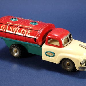 May include: A vintage-style toy tanker truck featuring a red cylindrical tank with the word GASOLINE printed in white. The truck has a cream-colored hood, a red cab roof, and a teal chassis. It includes black wheels and a silver-colored bumper.