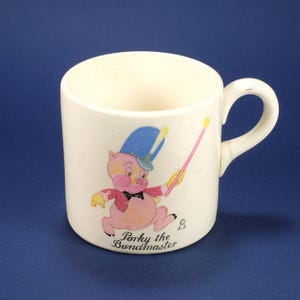May include: Cream ceramic mug with a cartoon image of Porky Pig as a bandmaster. The design includes a blue hat, pink jacket, and the text "Porky the Bandmaster". The mug has a curved handle.