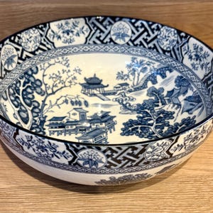 Może przedstawiać: Okrągła, biała ceramiczna miska z niebiesko-białym wzorem inspirowanym Azją. Miska przedstawia szczegółową scenę krajobrazu z pagodami, drzewami i mostami. Obręcz ma geometryczny wzór. Ta dekoracyjna miska nadaje się do serwowania lub ekspozycji.