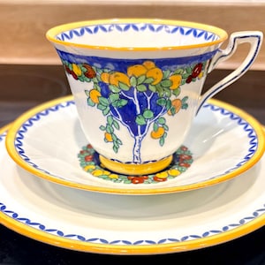 May include: A vintage teacup, saucer, and plate set. The cup and saucer feature a blue tree design with orange fruit and green leaves. The rims are trimmed with blue and yellow accents. The set is on a black surface.