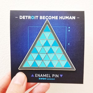 Op de afbeelding: Een blauwe en zilveren emaille pin met een driehoek die is opgebouwd uit kleinere driehoeken. De pin heeft de tekst "DETROIT BECOME HUMAN" en "ENAMEL PIN" erop.