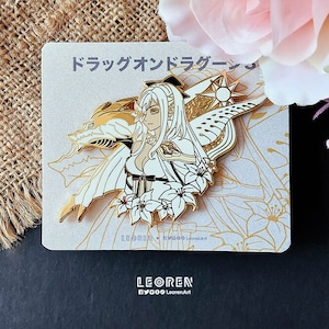Pode incluir: Um broche de esmalte dourado com uma mulher de cabelo branco com asas, um dragão e flores. O broche tem um fundo branco e detalhes dourados. O texto "ドラゴンライダー" está escrito em japonês no broche.