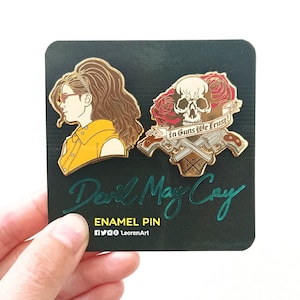 May include: Two gold enamel pins featuring characters from the video game Devil May Cry. The first pin is a portrait of a woman with brown hair and yellow clothing. The second pin features a skull with crossed pistols and the text "In Guns We Trust!"