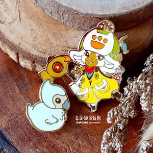 May include: Two enamel pins featuring cartoon characters. One pin depicts a character in a yellow outfit with a white hat and a red scarf, holding a golden object. The other pin is a light blue duck with a gold outline. The pins are on a wooden surface.