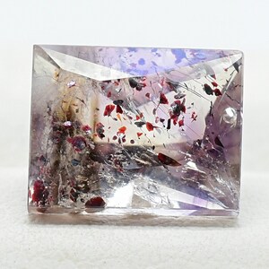 Puede incluir: Una gema de corte cuadrado con una superficie clara y multifacética. La piedra presenta inclusiones internas de minerales rojos, negros y marrones, creando un patrón único. La gema tiene un tono púrpura claro con áreas de transparencia.